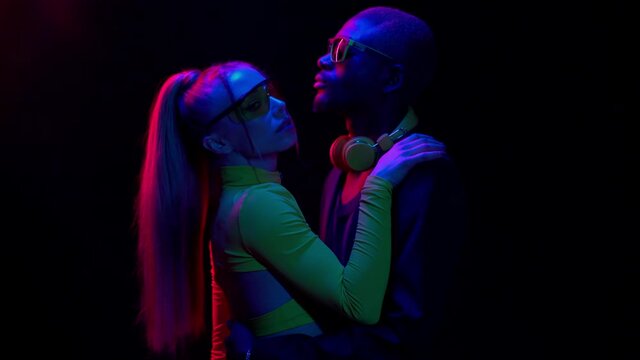 Interracial couple in neon lights standing close to each other. Happy couple in disco or lounge bar counter dancing alone on black background. Young people are having fun in the club.