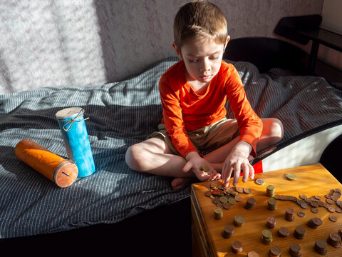 5-year-old Boy Puts Euro Cent Coins In His Palm,sits On Bed,selfmade Jar Banks Near Him,wooden Box With Scattered Coins Columns,sunny Day,hard Light.Learning Financial Literacy,savings.Selective Focus