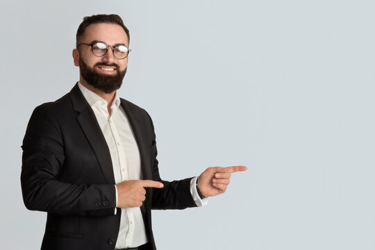 Serious businessman in office wear tand glasses smiling and pointing aside on light grey background, space for you ad
