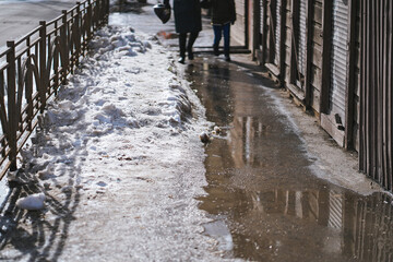Dirty muddy sidewalk with melting snow and puddles and water dripping from roofs. Public utilities problems in suburb and urban life. Snow removal, snow clearance