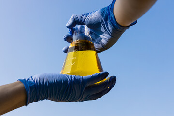 Hand with gloves holding beaker with ethanol biofuel against blue sky - obrazy, fototapety, plakaty