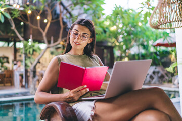Confident young ethnic woman reading planner while sitting at poolside with netbook