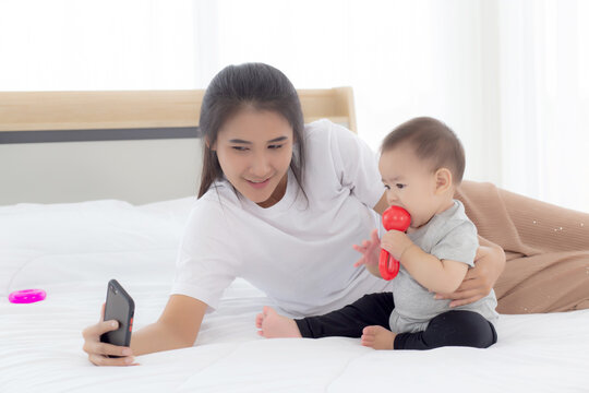 Young Asian Mother And Little Baby Girl Or Newborn Selfie With Smart Phone On Bed In Bedroom, Happiness Mom And Daughter Using Phone Video Call At Home, Two People, Family And Communication Concept.