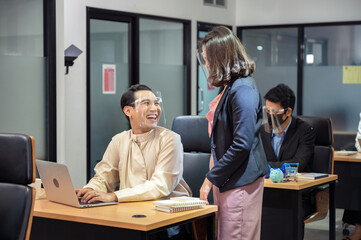 Asian businesswoman consulting with happy and smiling colleague wearing face shield at desk in modern office