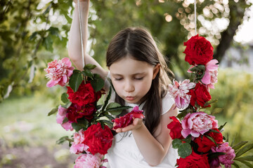 Portrait of a little girl 5 years old on a swing, decorated with flowers, roses and peonies. Girl blowing on flower petals. Spring and summer concept