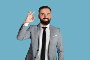 Cheerful young businessman in formalwear showing okay gesture on blue studio background