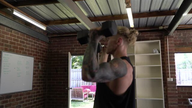 Tattoo man muscles in home gym doing dumbbell arnold press shot from side