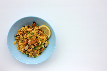 Traditional Spanish seafood dish paella with lemon slice on table. Mussel risotto with green peas on blue plate on white background. Top view, copy space. Healthy eating concept
