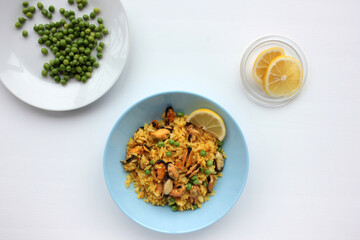 Traditional Spanish seafood dish paella with ingredients on table. Mussel risotto with green peas and lemon slice  on blue plate on white background. Top view, copy space. Healthy eating concept
