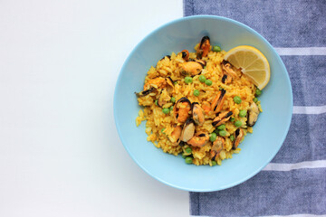 Traditional Spanish seafood dish paella with lemon slice on table. Mussel risotto with green peas in blue bowl on striped cloth napkin background. Top view, copy space. Healthy eating concept
