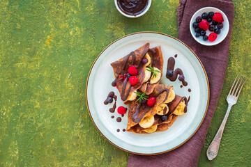 Thin chocolate pancakes stuffed with bananas, raspberries and blueberries, drizzled with chocolate sauce, on a gray ceramic plate on a green concrete background. Pancake recipes.