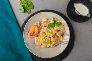 Farfalle pasta with shrimps in a creamy sauce on a gray plate on a concrete background next to a blue napkin and a jug of cream.