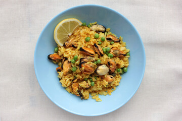 Traditional Spanish seafood dish paella with lemon slice on table. Mussel risotto with green peas on blue plate on white background. Top view, copy space. Healthy eating concept
