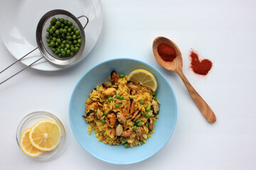 Traditional Spanish seafood dish paella with ingredients, lemon slice and wooden spoon with red paprika powder on table. Mussel risotto with green peas on blue plate on white background. Top view.