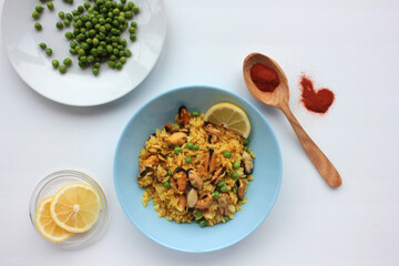 Traditional Spanish seafood dish paella with ingredients, lemon slice and wooden spoon with red paprika powder on table. Mussel risotto with green peas on blue plate on white background. Top view.