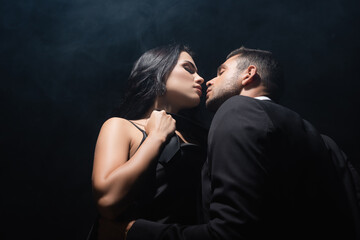 Low angle view of man kissing seductive woman in dress on black background with smoke