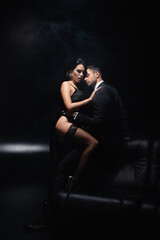 Man in suit hugging sensual woman in dress on couch on black background with smoke