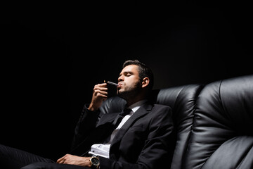 Man in suit holding lighter near cigarette on couch isolated on black