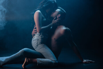 Sexy woman in jeans and bra kissing boyfriend on black background with smoke