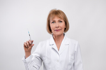 Portrait of mature female doctor in lab coat holding syringe with drug, vaccine or beauty injection on light background