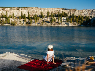 A traveler wearing a hat sits on a red cloth near the sea in nature