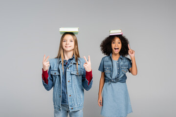 excited african american schoolgirl pointing with finger near happy friend showing victory signs isolated on grey