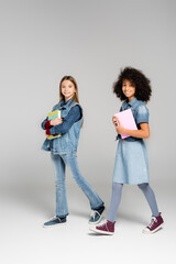 Fototapeta premium trendy interracial schoolgirls in denim clothes and gumshoes walking with textbooks on grey