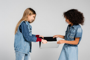 laughing interracial kids in denim clothes taking laptop from each other isolated on grey