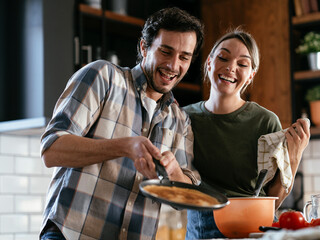Loving couple having fun while preparing food. Boyfriend and girlfriend making pancakes at home..