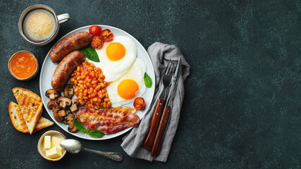 Full English breakfast on a plate with fried eggs, sausages, bacon, beans, toasts and coffee on dark stone background. With copy space. Top view