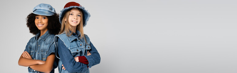 happy interracial girls in denim clothes and hats posing with crossed arms isolated on grey, banner