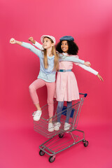 Obraz na płótnie Canvas fashionable, happy interracial girls posing in shopping cart on pink