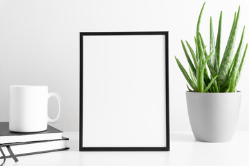 Black frame mockup with workspace accessories, mug of tea and aloe vera in pot on white table. Front view. Place for text, copy space, mockup