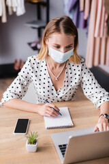 proprietor in medical mask, working on laptop near smartphone and notebook in showroom