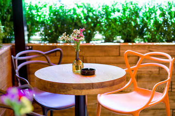 a round wooden table with two colored chairs and a glass vase with flowers on the countertop of a street cafe with bushes, empty seats, nobody.