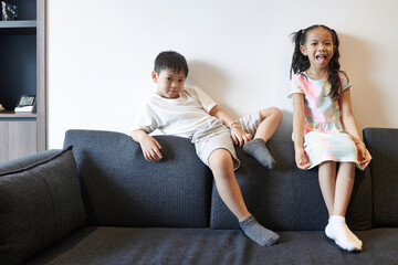 Funny preteen Vietnamese brother and sister sitting on sofa in living room and looking at camera