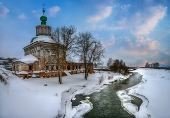 A view of the ancient unrestored stone church in the old Ural city of Solikamsk (Russia) on the banks of the Usolka River on a cold winter evening. Tall linden trees, white pure snow 