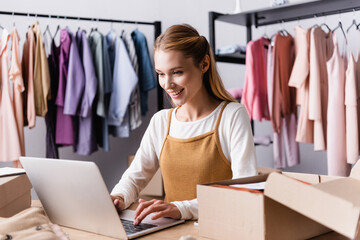 smiling showroom proprietor typing on laptop near cardboard boxes and racks on blurred background