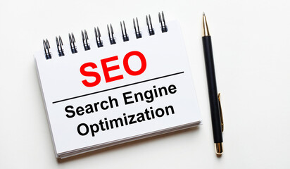 On a light background, a white notebook with are words SEO Search Engine Optimization and a pen.