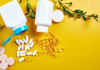 Flat lay of pills, fish oil, vitamins with green leavws on yellow background