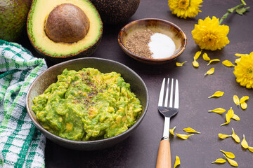 Fresh guacamole on a dish placed on a dark stone background with ingredients for homemade guacamole: avocados, lemon, salt, and pepper. Top view.  Concept of traditional Mexican preparation