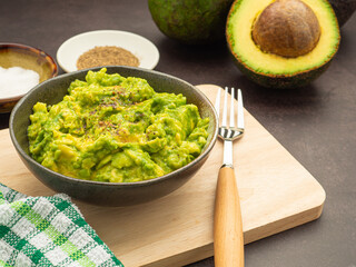 Fresh guacamole on a dish placed on a cutting wooden board with ingredients for homemade guacamole: avocados, lemon, salt, and pepper. Top view.  Concept of traditional Mexican preparation