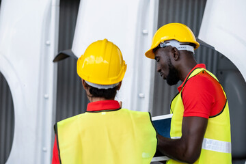 Two African America engineer wearing safety helmet and vest standing in the automotive warehouse with blur background. Man holding tablet and discuss with woman. Distribution and inventory concept