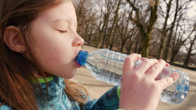 Little girl outdoors in the park drinks clean drinking water from a plastic bottle