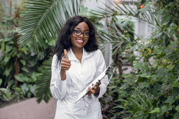 Young smiling African woman, professional scientist agronomist posing to camera with smile and thumb up, while standing in beautiful greenhouse with tropical plants.