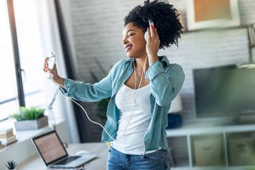 Motivated young afro woman listening to music with headphone while dancing in living room at home.