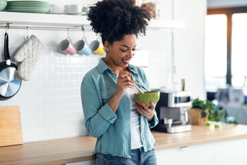 Beautiful afro woman eating noodles with chopsticks while standing in the kitchen at home.