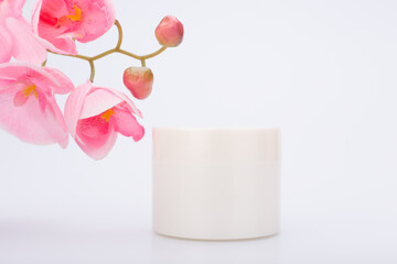 White glossy cream jar against white background with pink flower. Concept of natural or organic beauty products. Face mask, cream or scrub or hair mask for skin or hair care