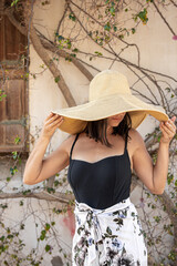 A girl in a large straw hat near a wall covered with a curly tree.