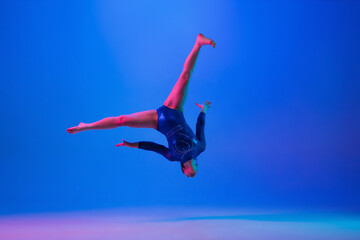 Jumping high. Young flexible girl isolated on blue studio background in neon light. Young female model practicing artistic gymnastics. Exercises for flexibility, balance. Grace in motion, sport